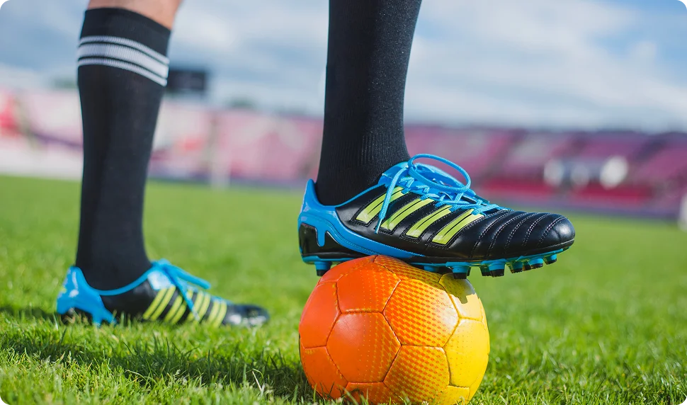 Un joueur de football en chaussettes noires et crampons rayés pose un pied sur un ballon orange, sur de l'herbe verte, avec un stade flou en arrière-plan sous un ciel dégagé.