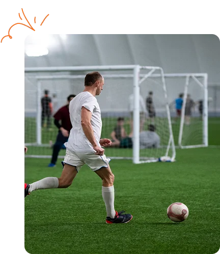 Trois hommes jouent au football en salle : l’un, en maillot blanc, dribble le ballon, poursuivi par deux autres en maillot rouge. L’ambiance est animée et compétitive.