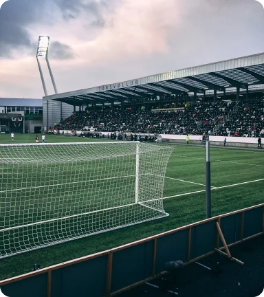 Un stade de football animé sous un ciel nuageux ; le regard est porté sur un poteau de but vide, avec une tribune de spectateurs bondée en arrière-plan.